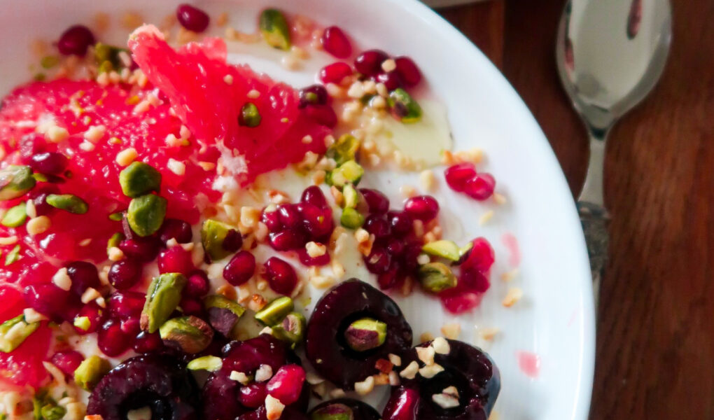 Overhead shot of a breakfast bowl with yoghurt, cherries, grapefruit, and pistachios layered in a ceramic dish