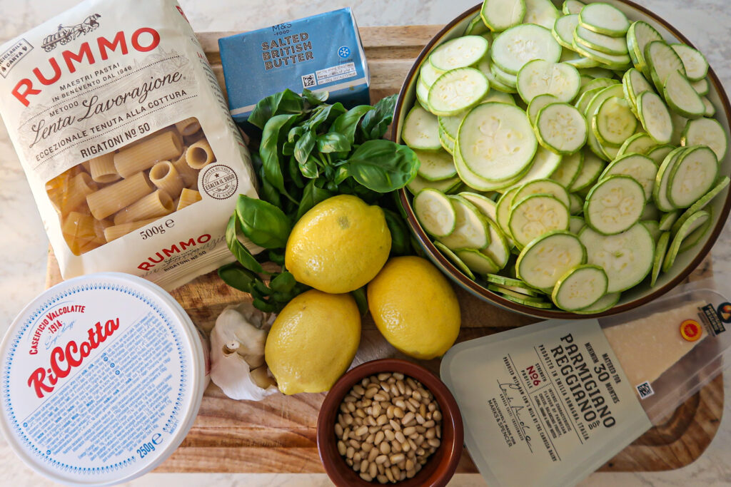 Overhead of ingredients for courgette lemon ricotta rigatoni: rigatoni, sliced courgettes, ricotta, double cream, lemons and zest, Parmesan, basil, garlic, olive oil, toasted pine nuts, salt and pepper.