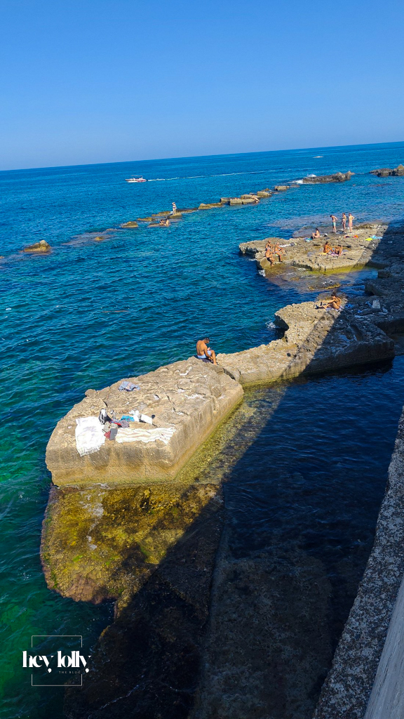 Wooden sunbathing platform with swimmers diving into clear turquoise waters in Ortigia