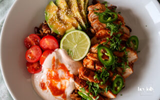 Chicken burrito bowl topped with sliced chicken, avocado, beans, sweetcorn, cheddar, and chilli cream drizzle.
