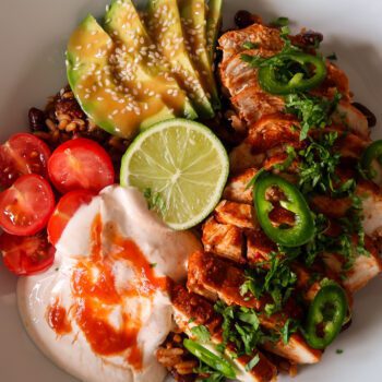 Chicken burrito bowl topped with sliced chicken, avocado, beans, sweetcorn, cheddar, and chilli cream drizzle.