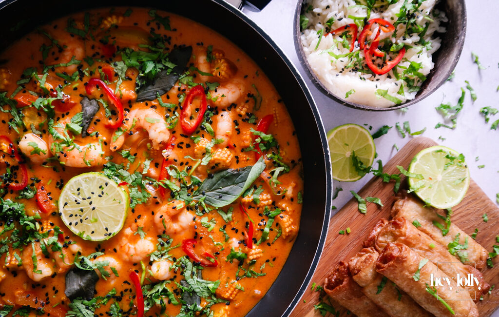 Overhead shot of fresh ingredients for Thai Red King Prawn curry including prawns, baby corn, red pepper, and courgette on a wooden board.