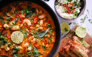 Overhead shot of fresh ingredients for Thai Red King Prawn curry including prawns, baby corn, red pepper, and courgette on a wooden board.