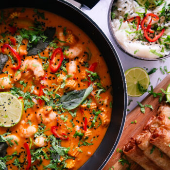 Overhead shot of fresh ingredients for Thai Red King Prawn curry including prawns, baby corn, red pepper, and courgette on a wooden board.
