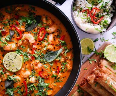 Overhead shot of fresh ingredients for Thai Red King Prawn curry including prawns, baby corn, red pepper, and courgette on a wooden board.