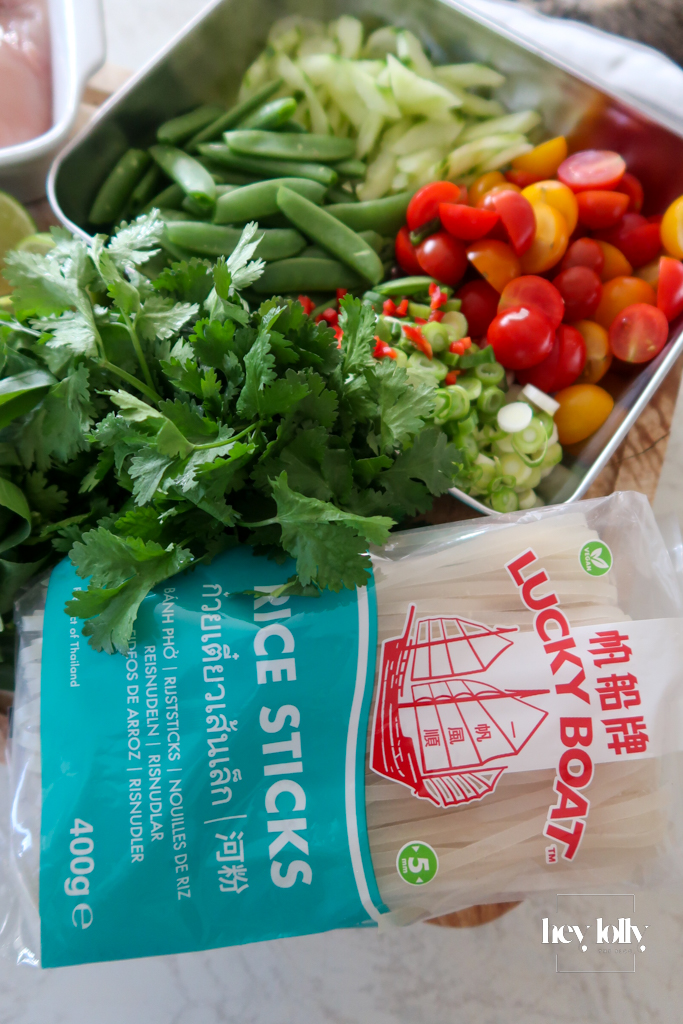 Fresh ingredients for Thai poached chicken salad laid out: glass noodles, cherry tomatoes, cucumber, sugar snap peas, herbs, lime, and coconut milk.