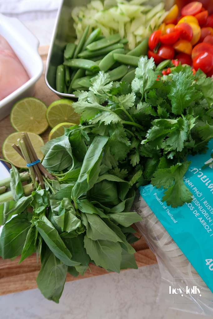 Fresh ingredients for Thai poached chicken salad laid out: glass noodles, cherry tomatoes, cucumber, sugar snap peas, herbs, lime, and coconut milk.