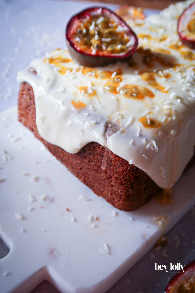 Passion fruit yoghurt loaf with cream cheese frosting on wooden board surrounded by cut passion fruits and serving knife.