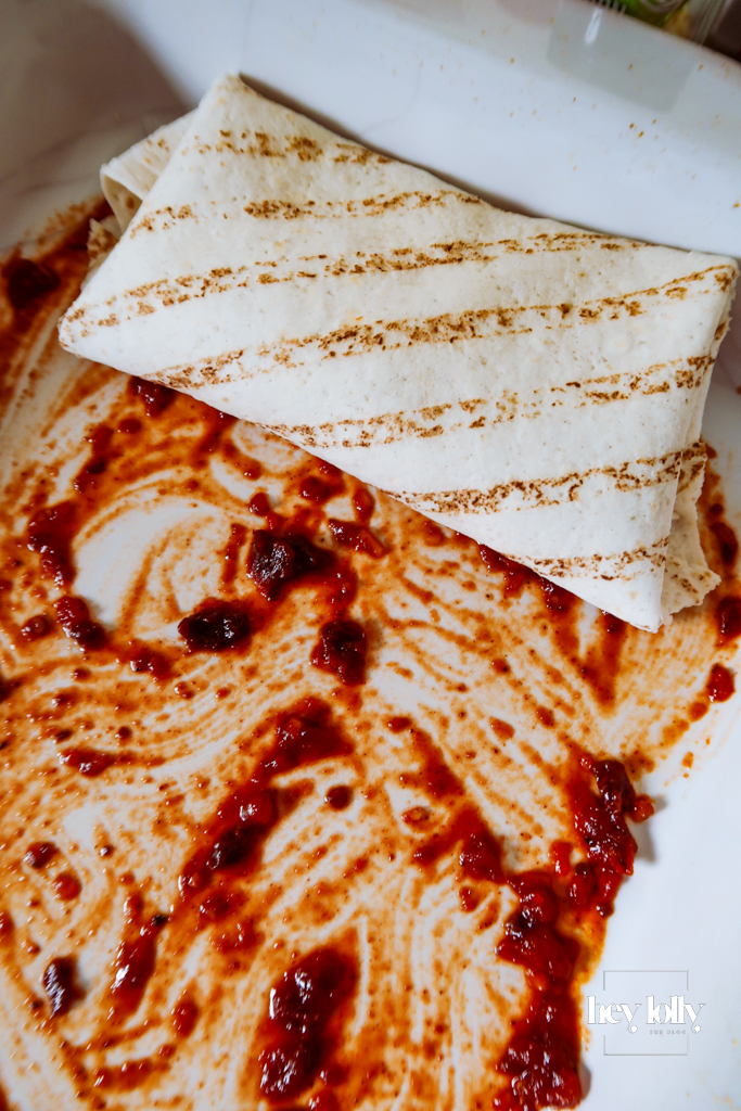 Rolling tortillas filled with corn and butter bean mixture on the worktop, sauce splatters visible around.