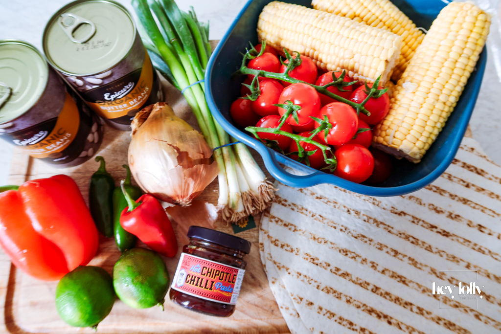 Flat lay of fresh ingredients for charred sweetcorn and butter bean enchiladas — corn cobs, red pepper, butter beans, chipotle paste, tortillas, lime and cheese on a rustic wooden board.