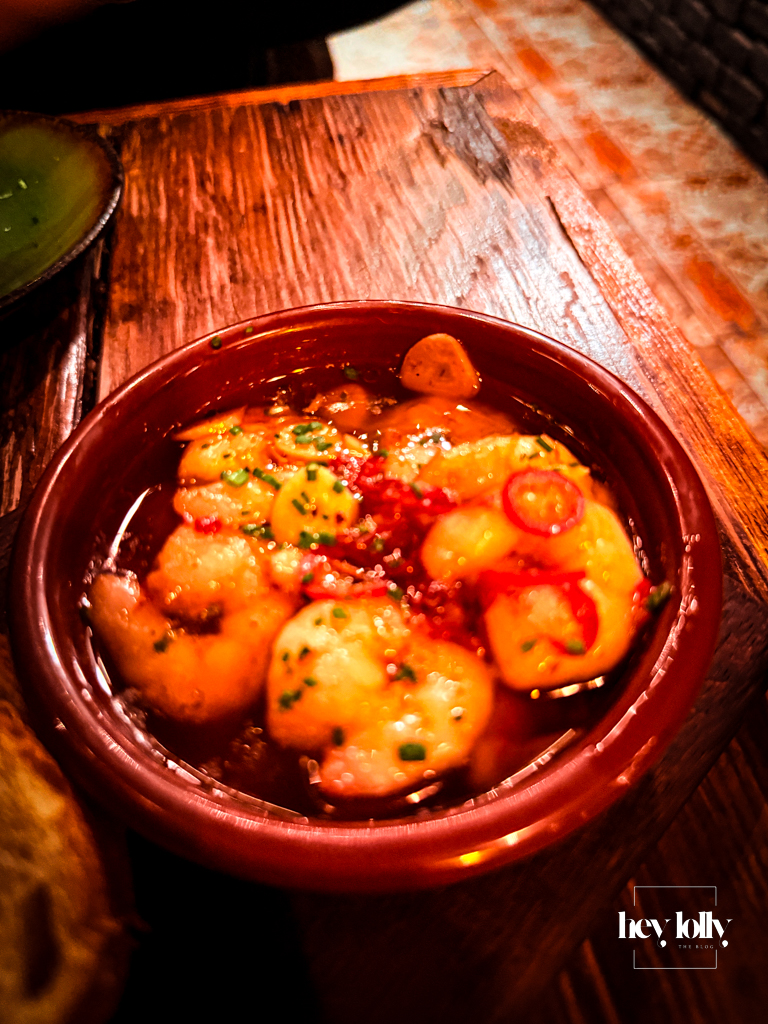 Gambas Pil Pil at Plates by Purnell’s – king prawns in garlicky chilli oil served in a terracotta dish on a wooden table.
