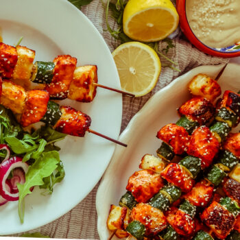 Salmon, Courgette & Halloumi Skewers with Lemon Tahini Dressing, topped with herbs and lemon wedges.