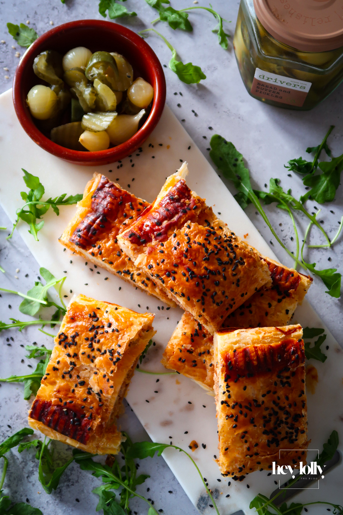 Golden sun-dried tomato chutney sausage rolls topped with black sesame, flaky puff pastry and melted Cheddar visible inside.