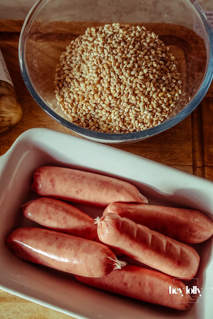 Base ingredients for pearl barley stew laid out on a wooden board, including sausages, carrots, onions, garlic, kale, white beans and herbs