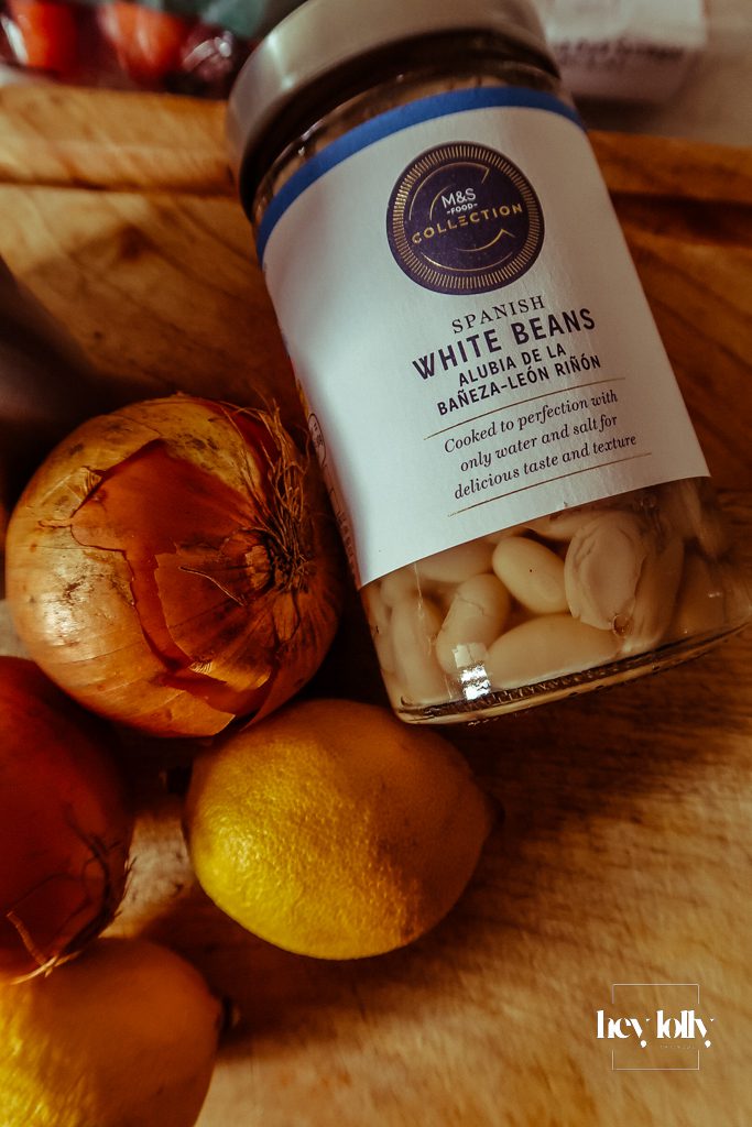 Base ingredients for pearl barley stew laid out on a wooden board, including sausages, carrots, onions, garlic, kale, white beans and herbs
