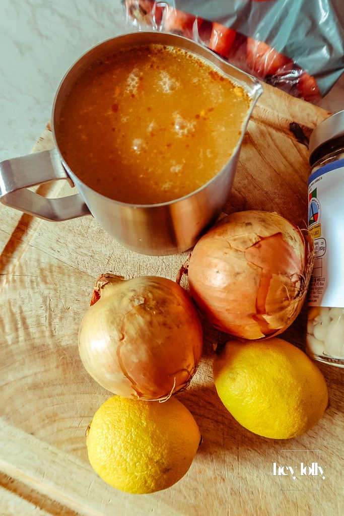 Base ingredients for pearl barley stew laid out on a wooden board, including sausages, carrots, onions, garlic, kale, white beans and herbs