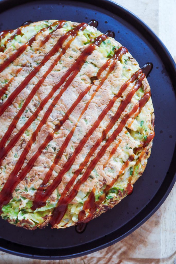 overhead shot of okonomiyaki pancake with gochujang, spring onions and sesame seeds on a white rustic table