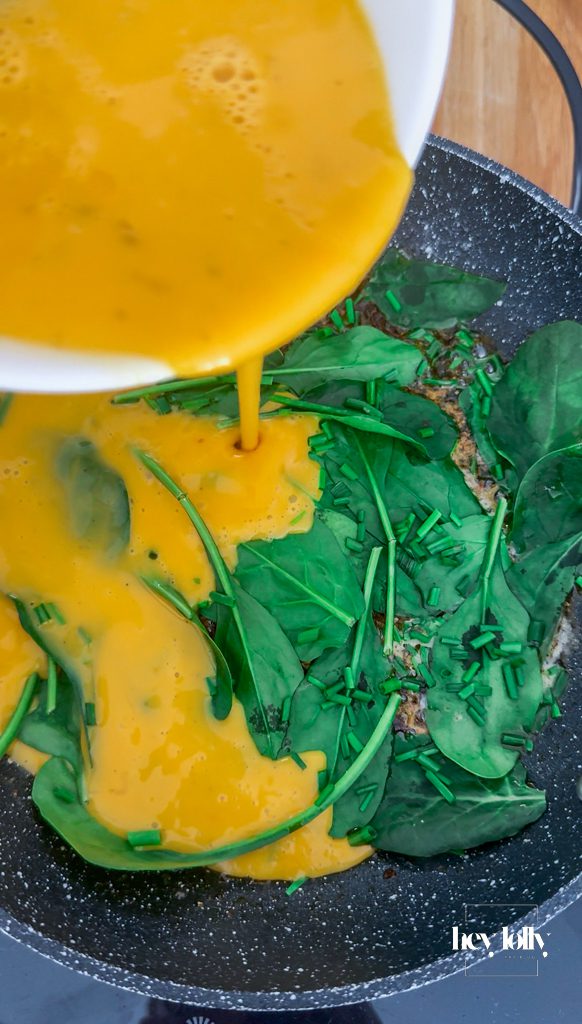 Egg mixture added to spinach, chives and spring onions wilting in pan with chilli crisp for breakfast quesadilla